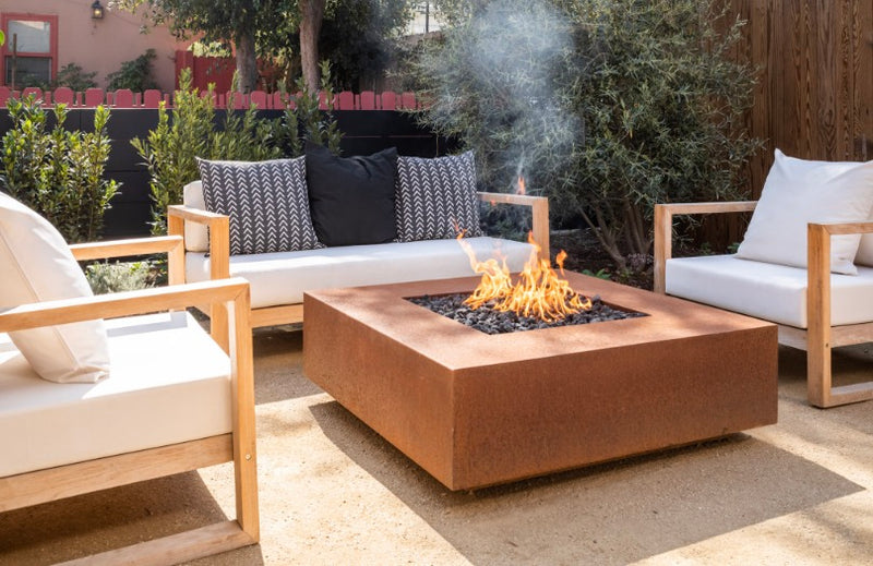 Corten steel square gas fire pit with teak lounge chairs on a gravel patio