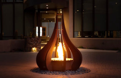 The Outdoor Plus Comet corten steel teardrop fire sculpture glowing at night outside a modern home