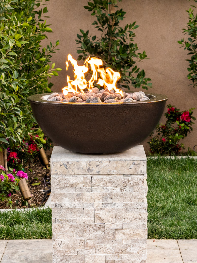 Dark round gas fire bowl on a travertine stone pedestal in a garden